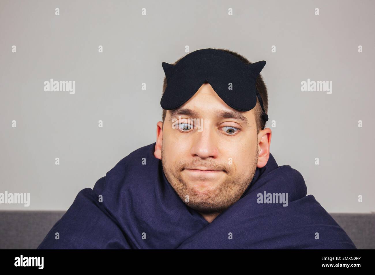 Embarrassed young man in a blanket at home wears a sleep mask while