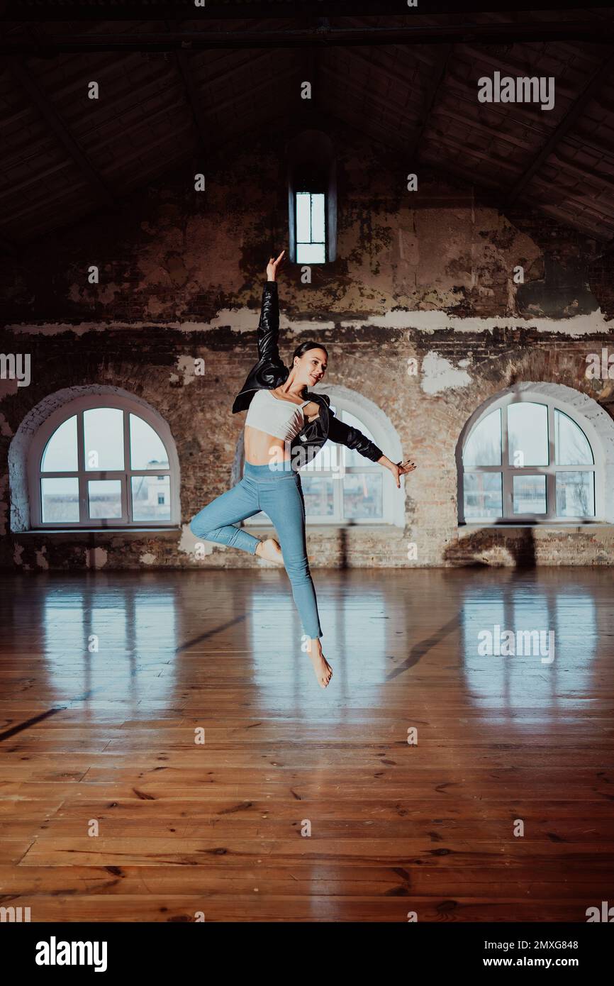 Young woman jumping in casual style - jeans and leather jacket doing ballet in old studio. Attractive ballerina practices in choreography alone. High Stock Photo