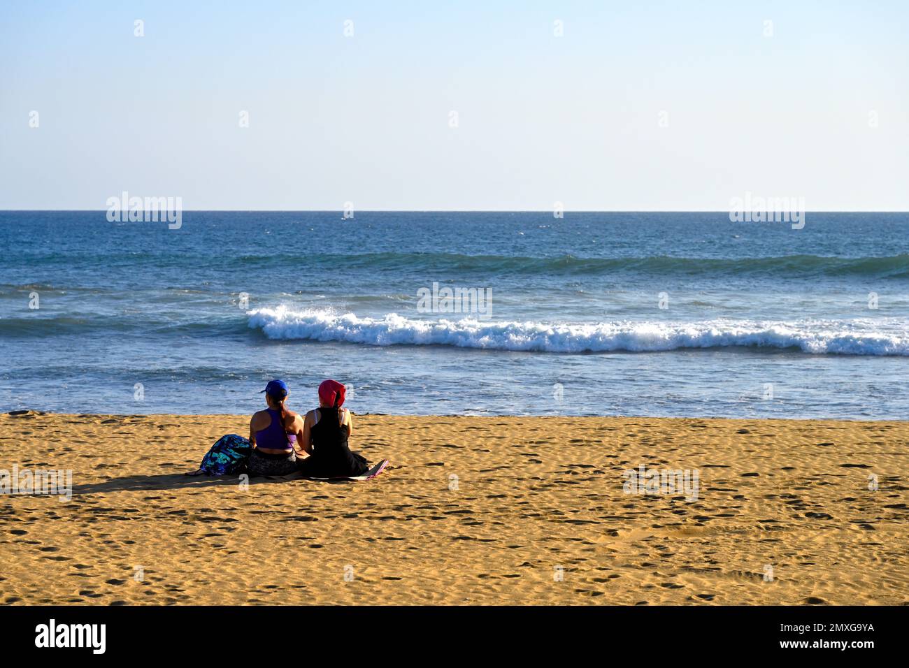 Two people sitting on sandy beach looking out to breaking waves at seaside, Maspalomas, Gran Canaria Stock Photo