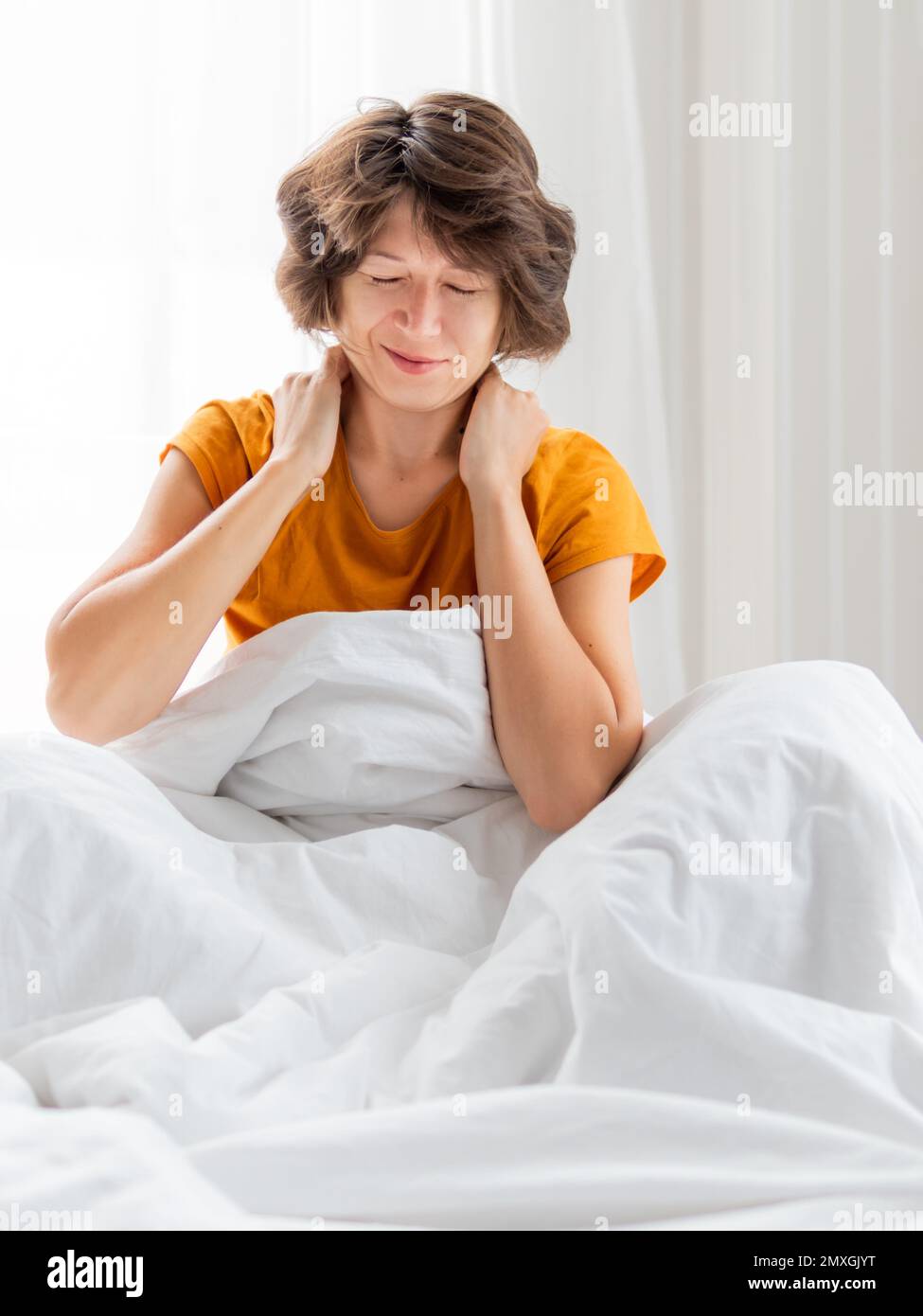 Sleepy woman in yellow pajama is massaging her neck. Waking up early in morning. Woman gets enough sleep. Stock Photo