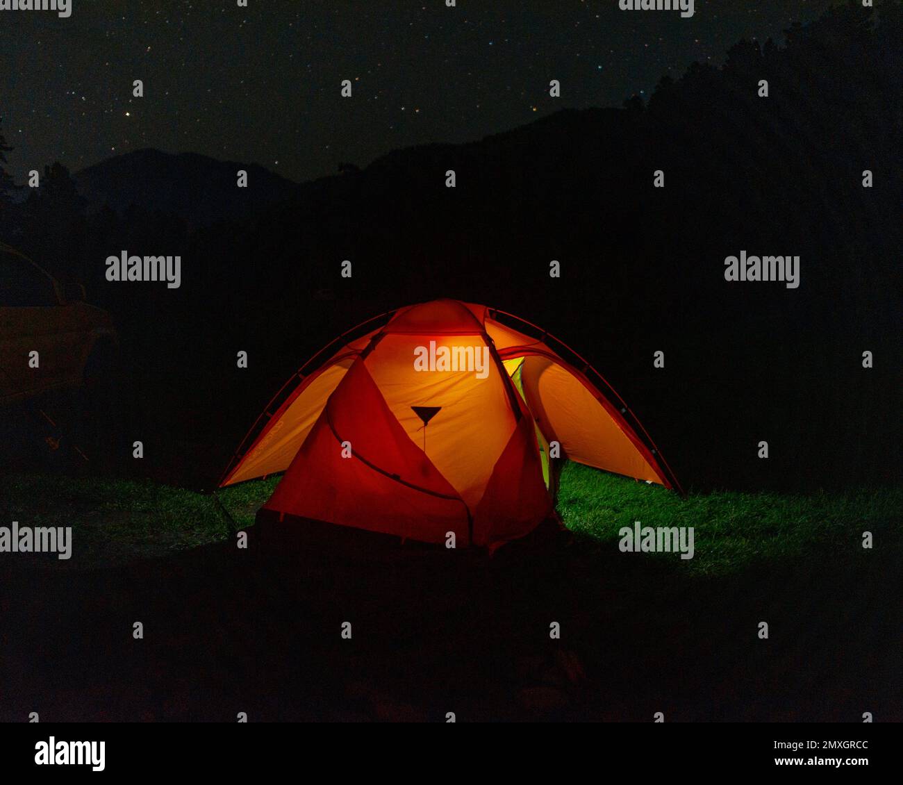 Tourist tent of travelers with an open entrance under the starry sky in the mountains covered with forest at night next to the edge of the car. Stock Photo