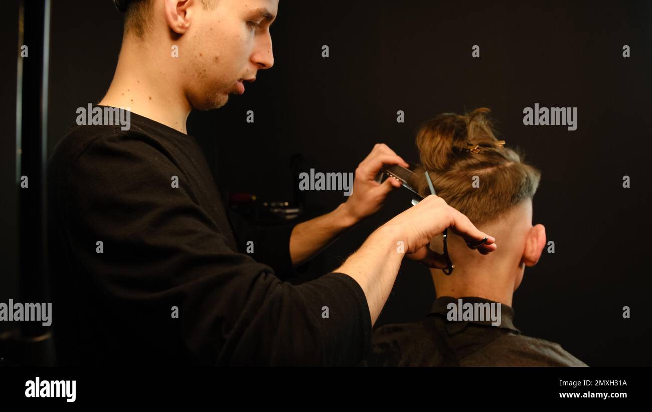 Barber use Thinning scissors and metal comb on brown wavy hair of young man. Hairdresser service in a modern barbershop in a dark key lightning with w Stock Photo