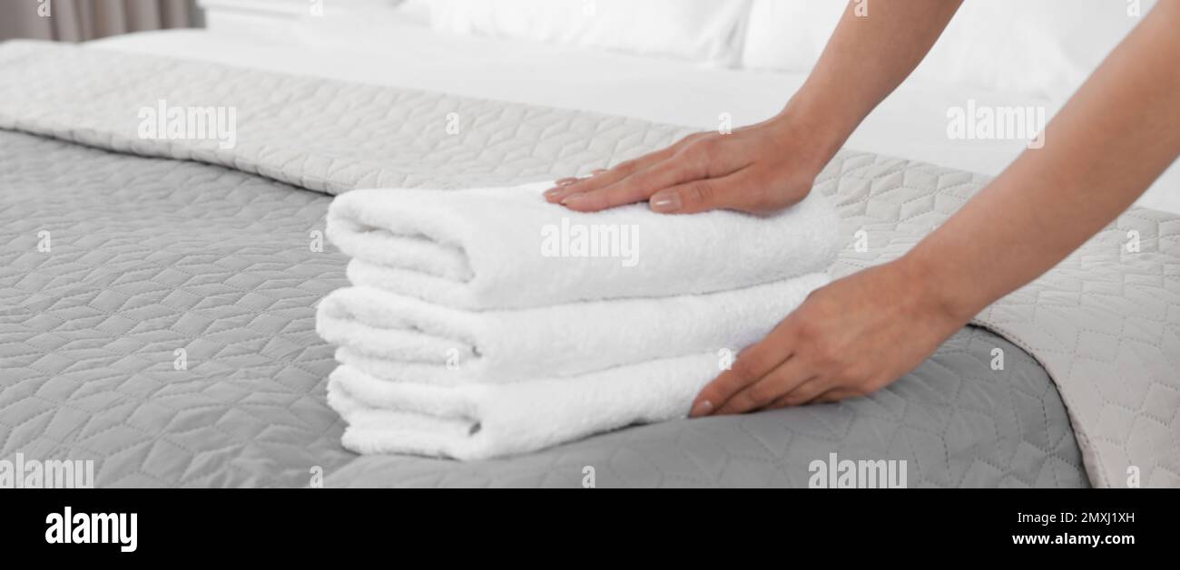 Young maid putting stack of fresh towels on bed in hotel room, closeup