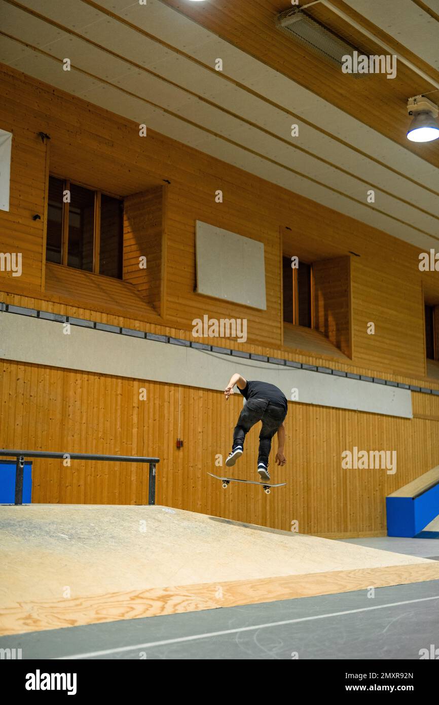 A vertical shot of a skater jumping over the skating ramp inside a building. Stock Photo