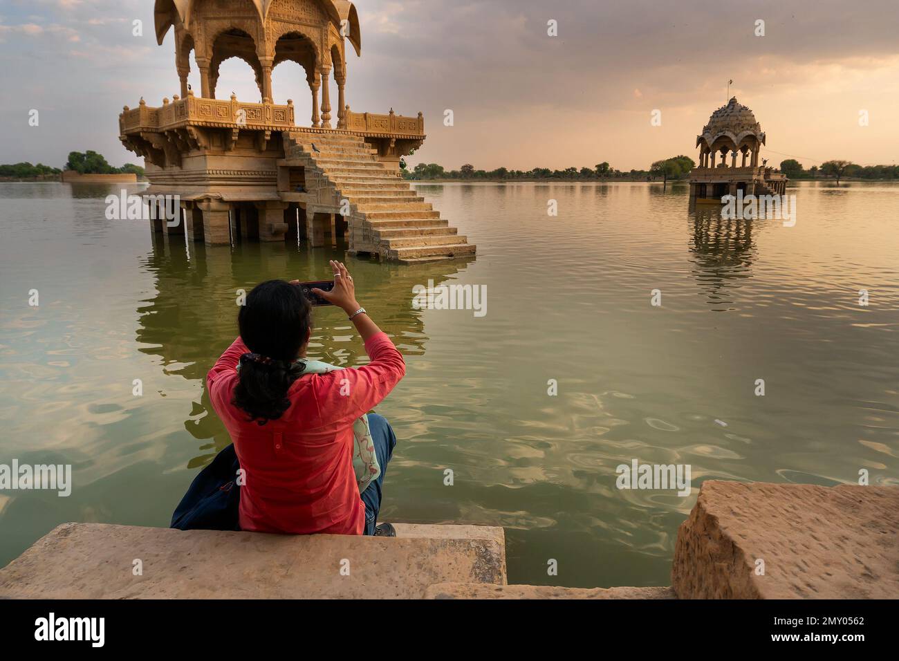 Indian female traveller, woman photographer taking picture of Chhatris and shrines with reflection of them on the water of Gadisar lake; Jaisalmer. Stock Photo