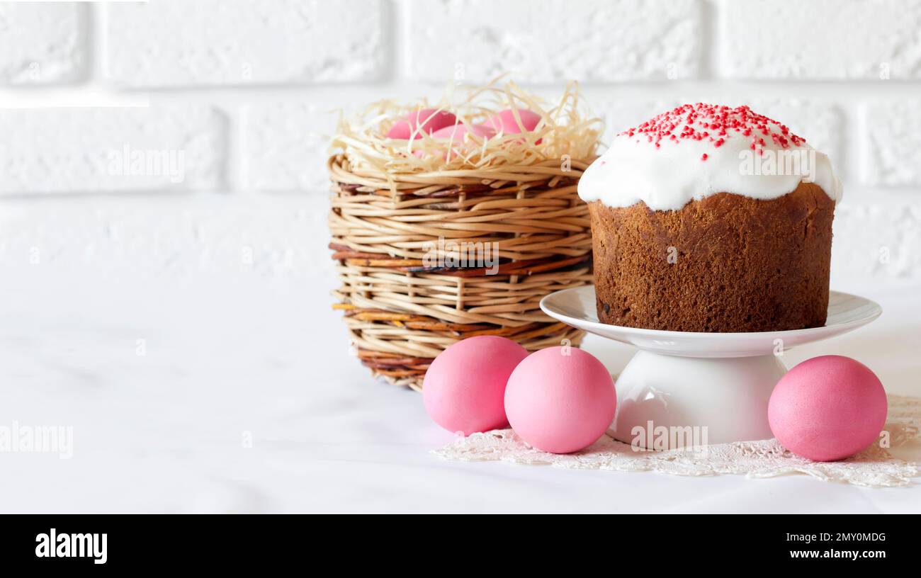 Minimalistic Easter composition with wicker basket with pink colored eggs and Easter cake on white background. Copy space Stock Photo
