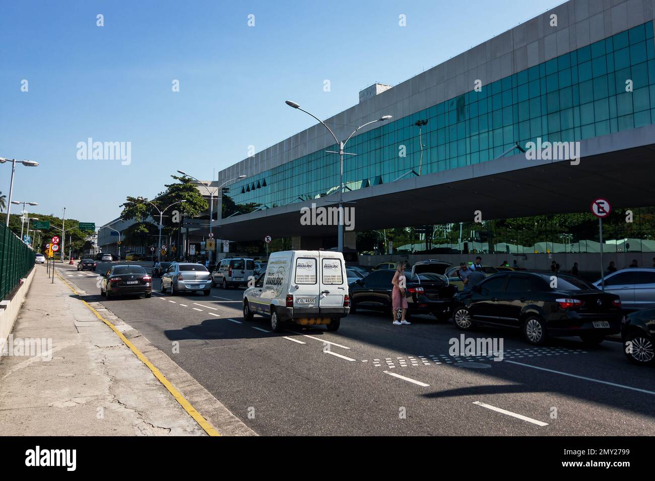 Lots of cars stopped at the populated outdoor departure area of Santos Dumont airport while traffic pass by nearby under summer sunny clear blue sky. Stock Photo