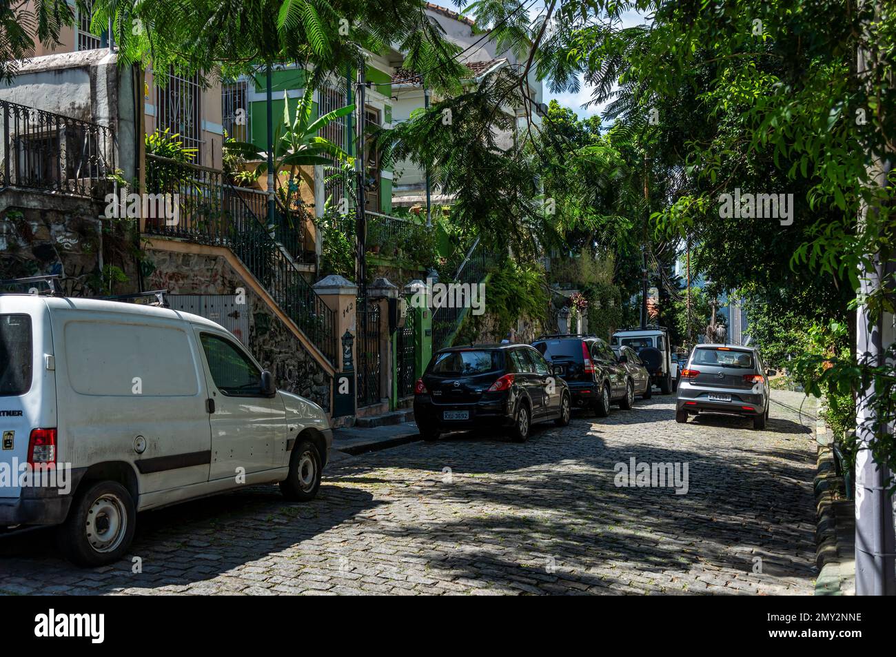 Cars parked on Ladeira de Santa Teresa cobblestone slope street in front of some houses in Santa Teresa district under summer afternoon sunny blue sky Stock Photo