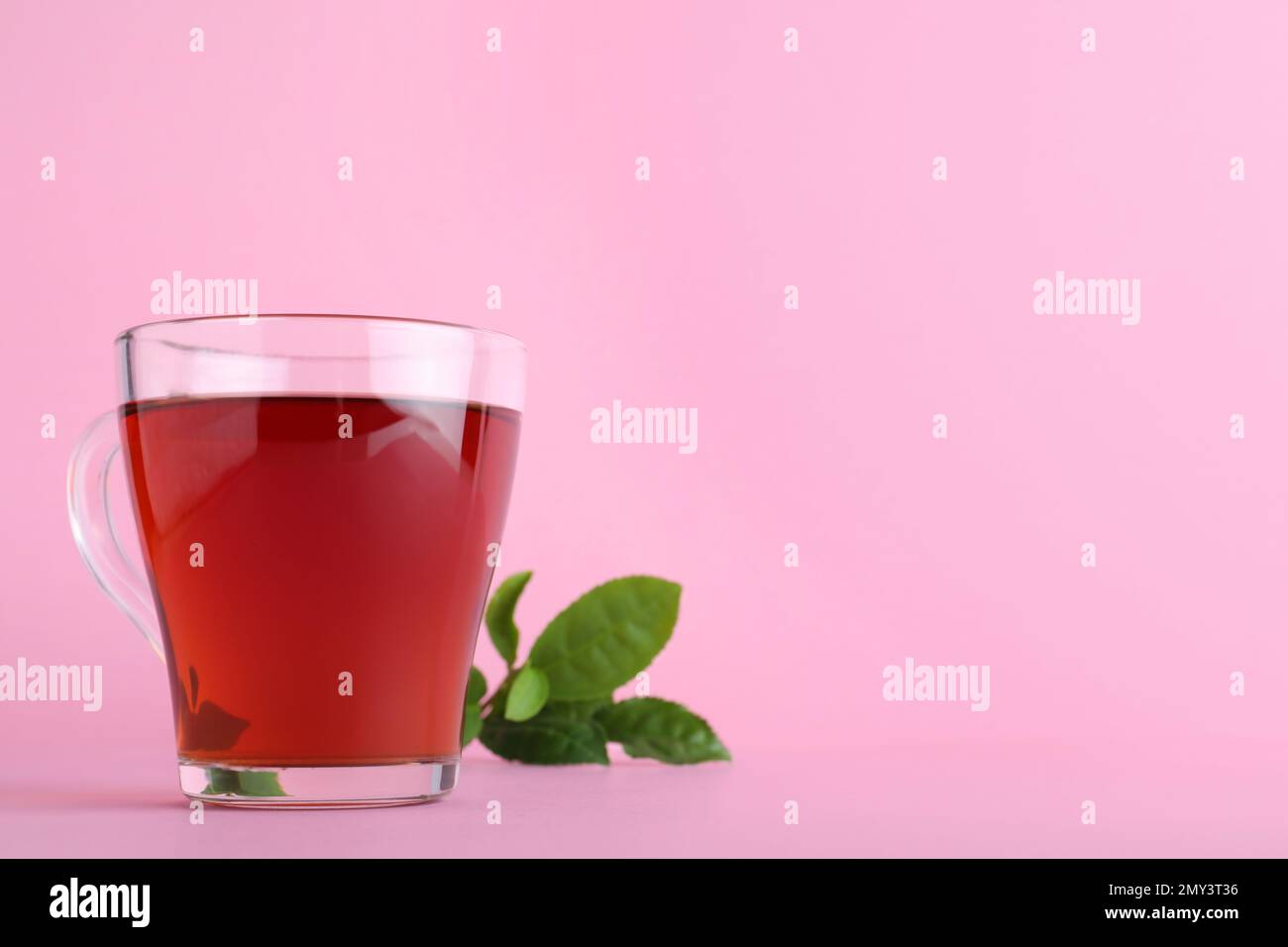 Glass cup of aromatic black tea and green leaves on pink background
