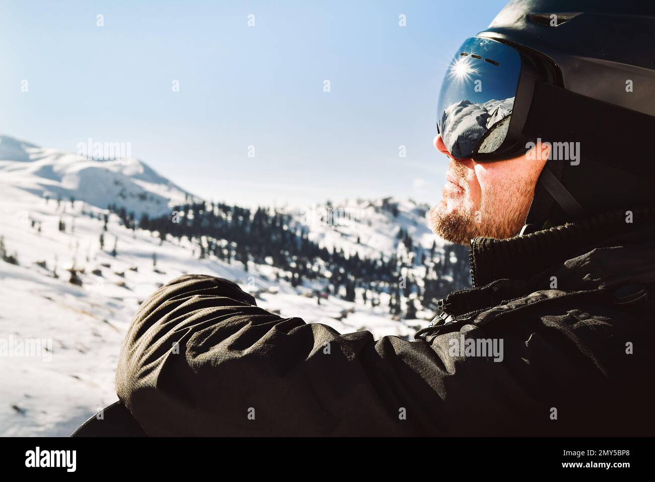 Close up of the ski goggles of caucasian man with the reflection of snowed mountains and sunburst. A mountain range reflected in the ski mask. Portrai Stock Photo