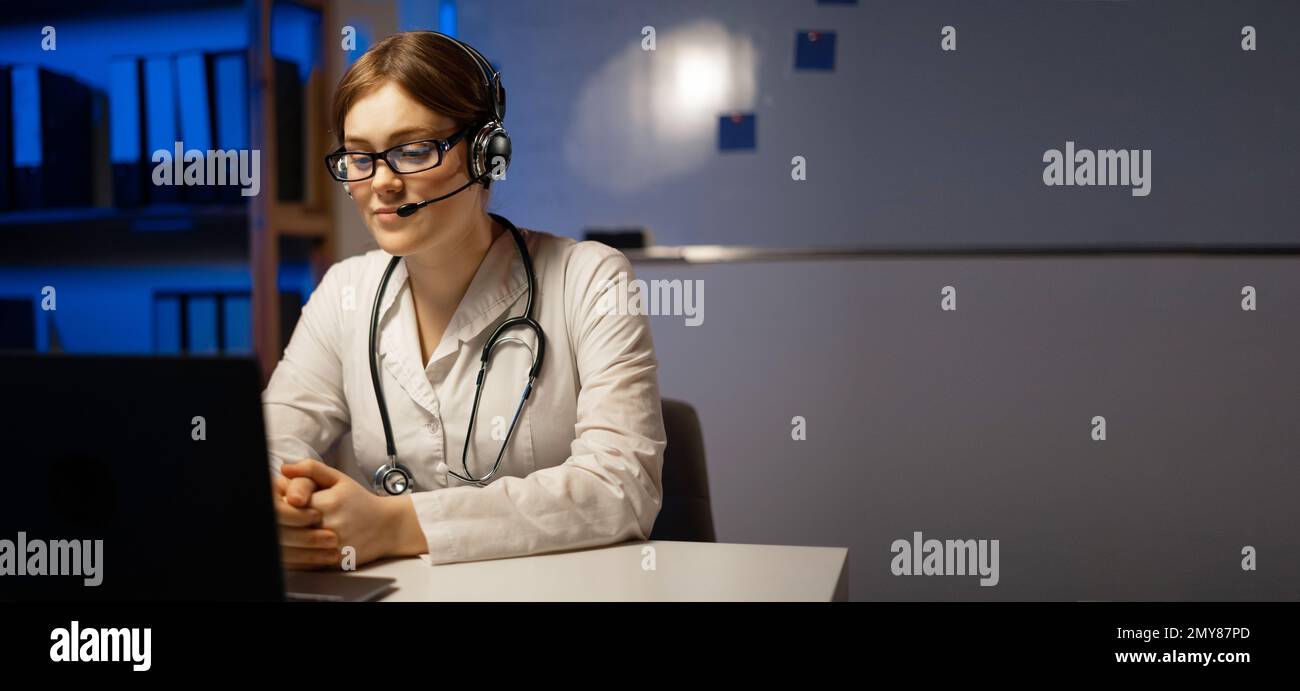 Attractive doctor wears a headset use laptop webcam for video call or conference, working at night shift. Remote online medical chat consultation, vir Stock Photo
