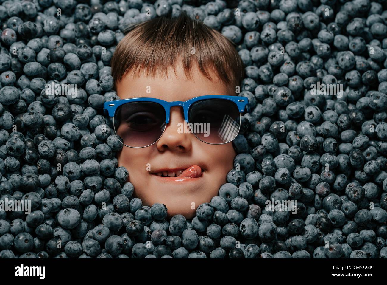 Smiling little boy face in eyewear fresh ripe berries - blueberries. Child covered with blackberry. Kid enjoying organic bilberry plant. Diet, antioxi Stock Photo