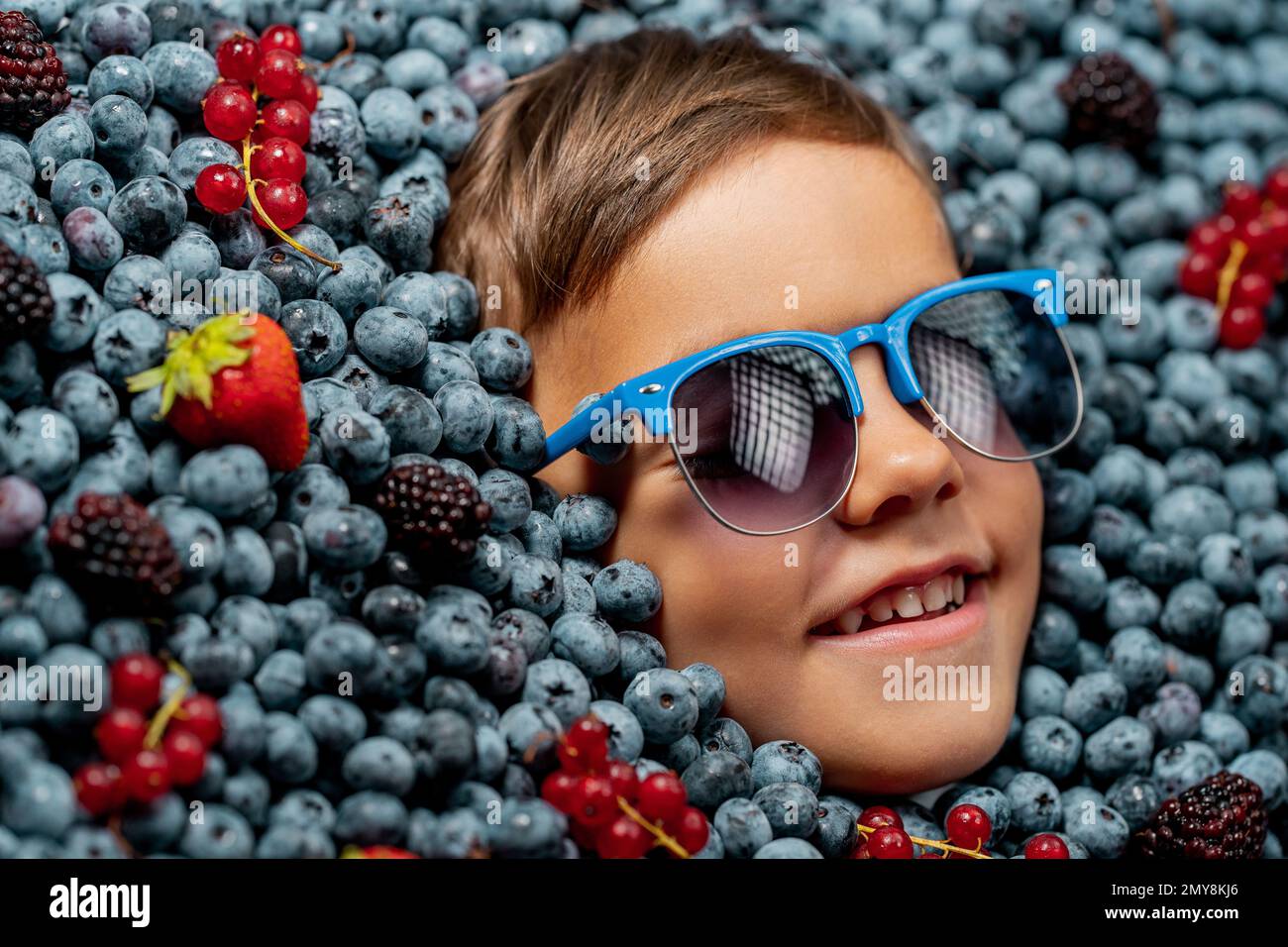 Smiling little boy face in eyewear fresh ripe berries - blueberries, strawberries, currant. Child covered with blackberry. Kid enjoying organic bilber Stock Photo