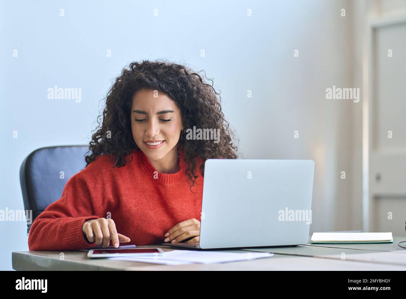 Young smiling latin business woman sitting at office desk using digital tablet. Stock Photo