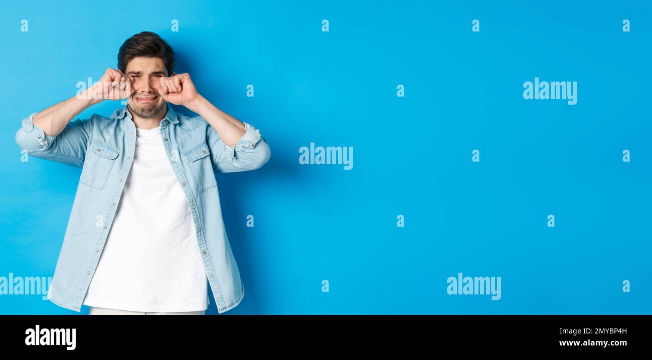 Sad timid man crying, wiping tears off face and sobbing, looking offended and upset, standing over blue background Stock Photo