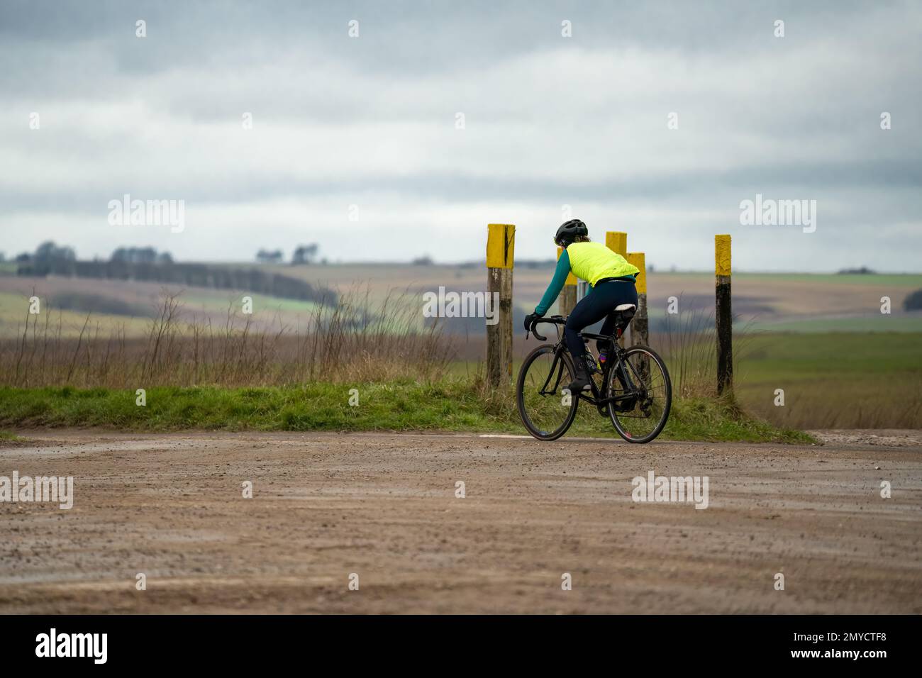 a sports cyclist wearing a yellow hig-vis top, riding a racing bike past tank crossing marker posts Stock Photo