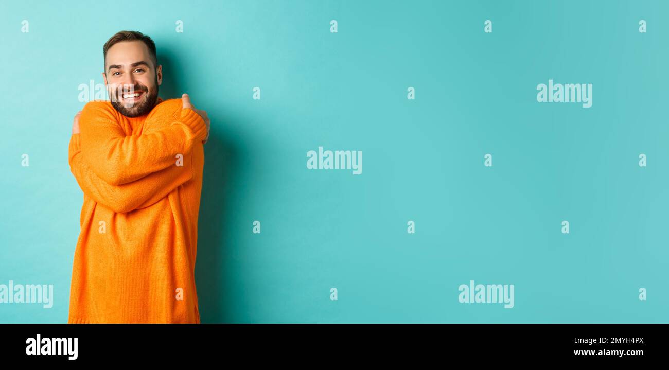 Happy man feeling comfortable, wearing warm sweater and hugging himself, smiling satisfied, standing over light blue background Stock Photo