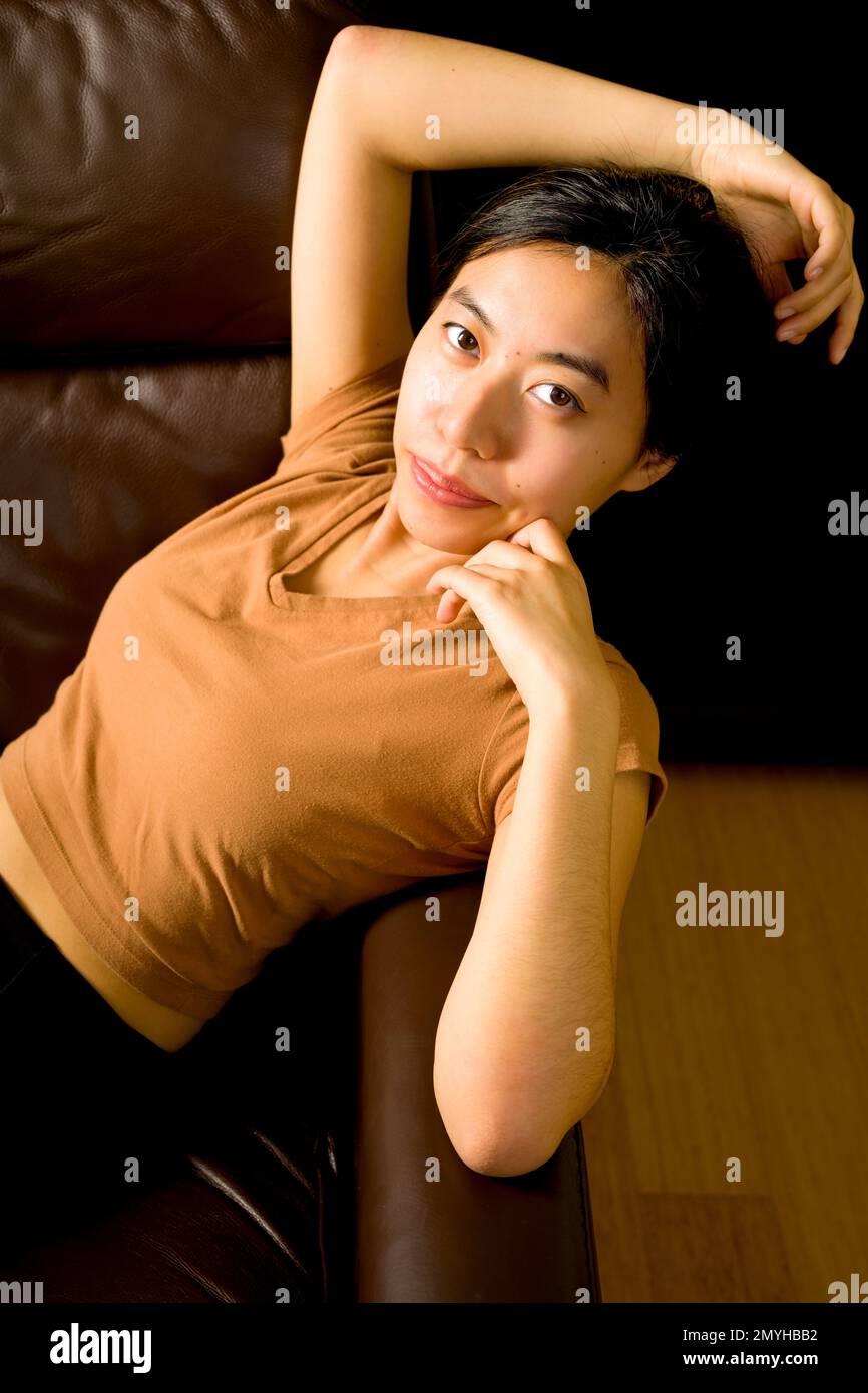 Beautiful Young Asian Woman Reclining on a Brown Leather Chair Against