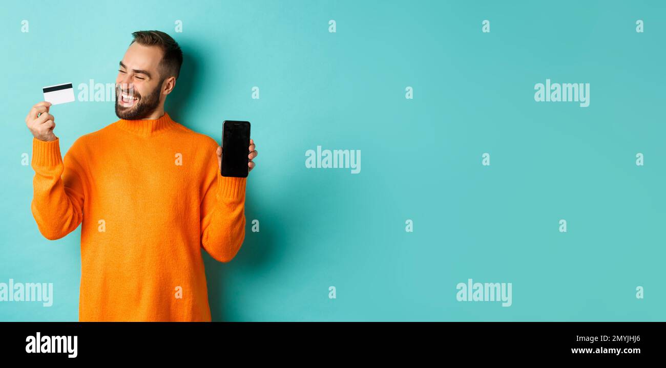 Online shopping. Satisfied man using credit card and showing mobile screen, looking pleased, standing over light blue background Stock Photo