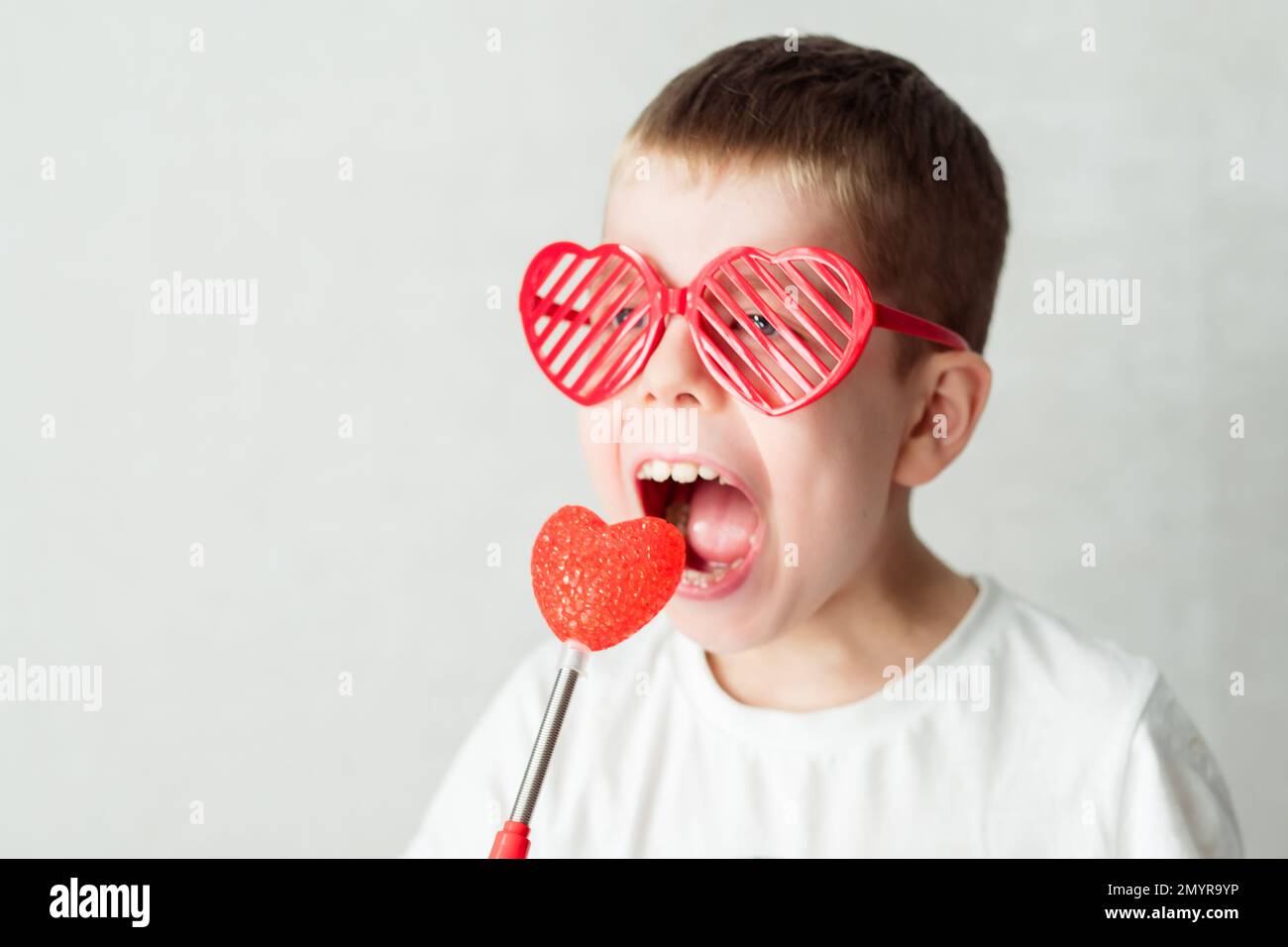 portrait of a boy in heart-shaped glasses in a white t-shirt against a white background biting a heart. Against valentine's day Stock Photo