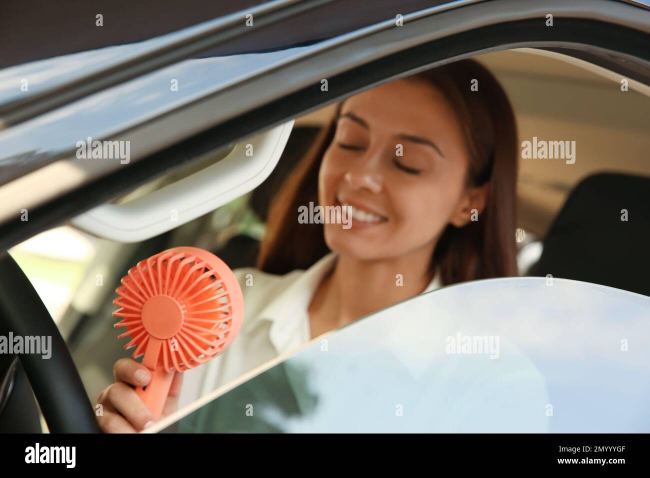 Young woman enjoying air flow from portable fan in car on hot summer