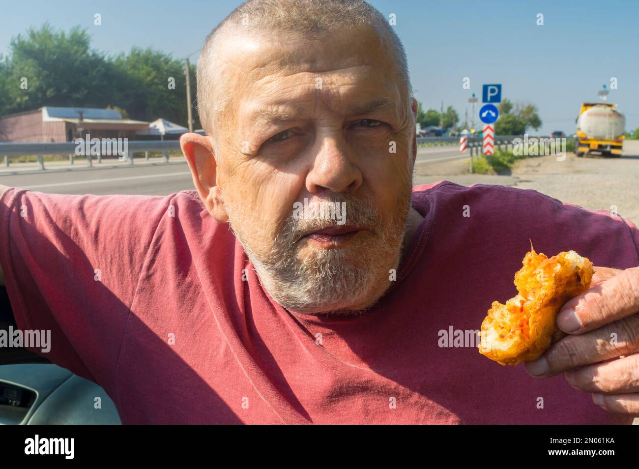 Portrait of hungry Ukrainian senior driver taking patty near his car while standing near Vasylivka, Zaporizhzhia Oblast, Ukraine Stock Photo