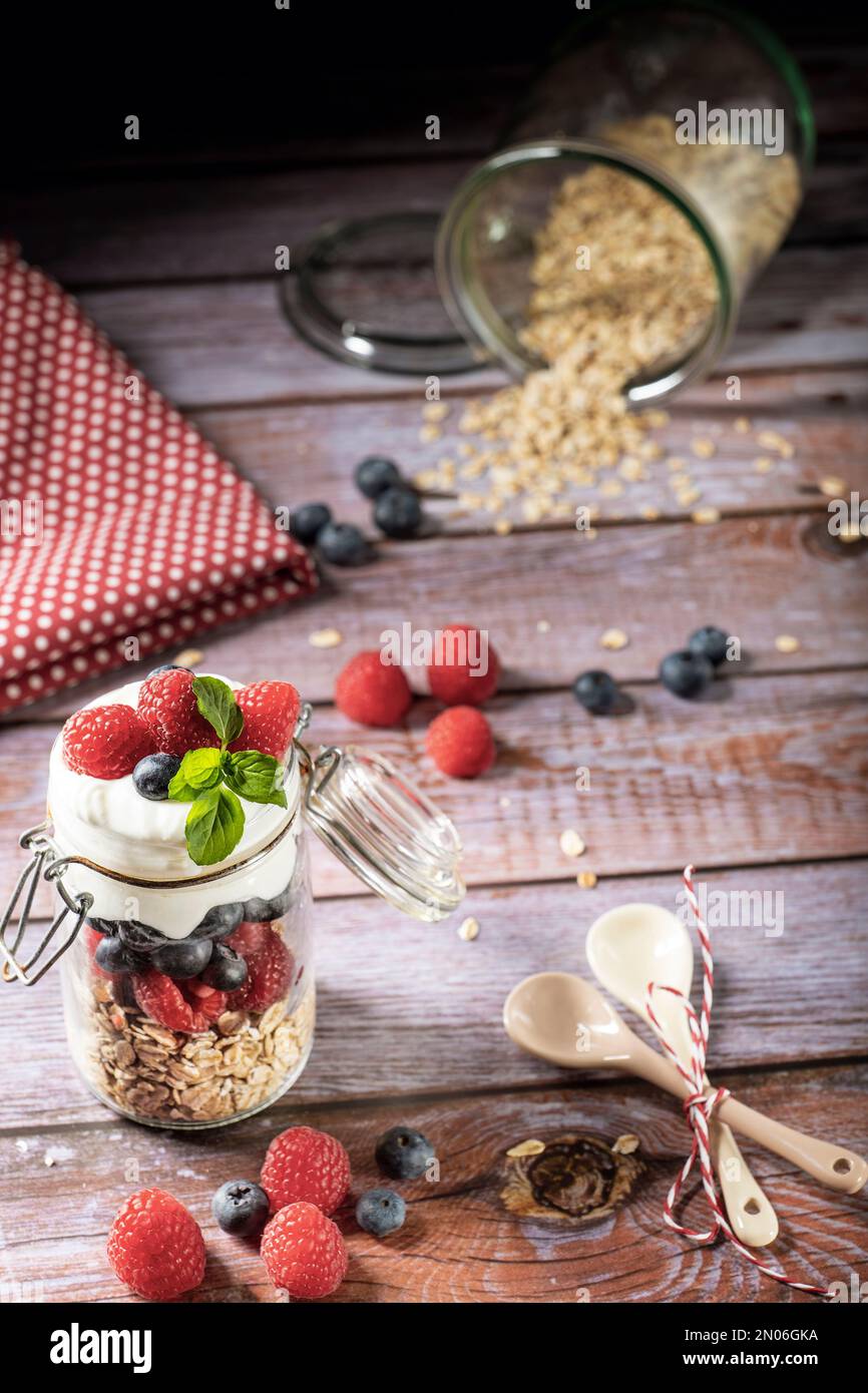 Muesli layered in a glass with oatmeal, raspberries, blueberries and yogurt. Decorated on a rustic table top with berries, spoons, towel. Stock Photo