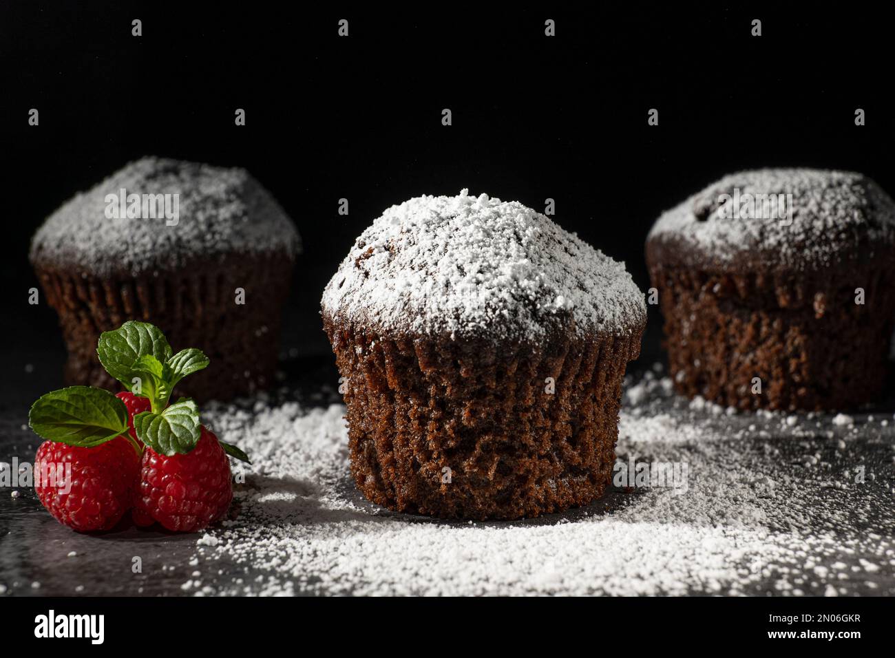 Low key shot of chocolate muffins sprinkled with powdered sugar. Close up, decorated with fresh yewberries and a sprig of mint Stock Photo