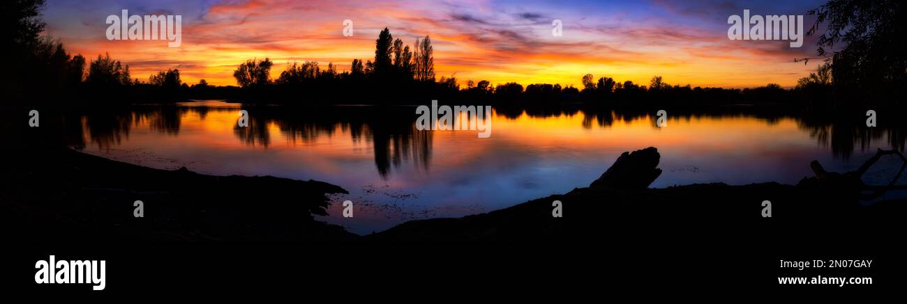 Magnificent colorful sunset at a lake, extra wide panorama with dark silhouettes of trees and the lakeshore, with the dramatic pretty sky reflected in Stock Photo