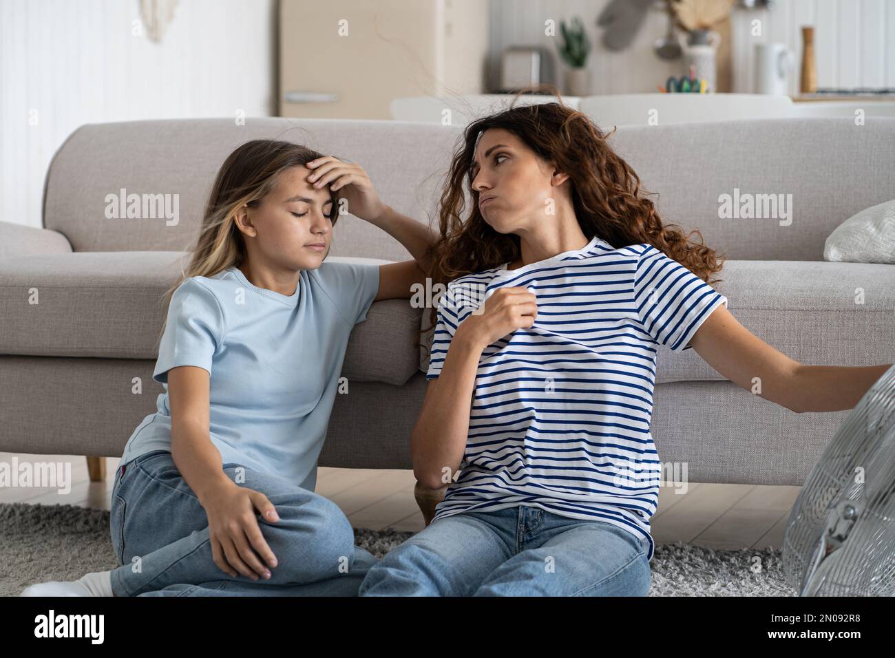 Sweaty tired woman sits on floor next to sofa and fan looking back at teenage girl after summer walk Stock Photo