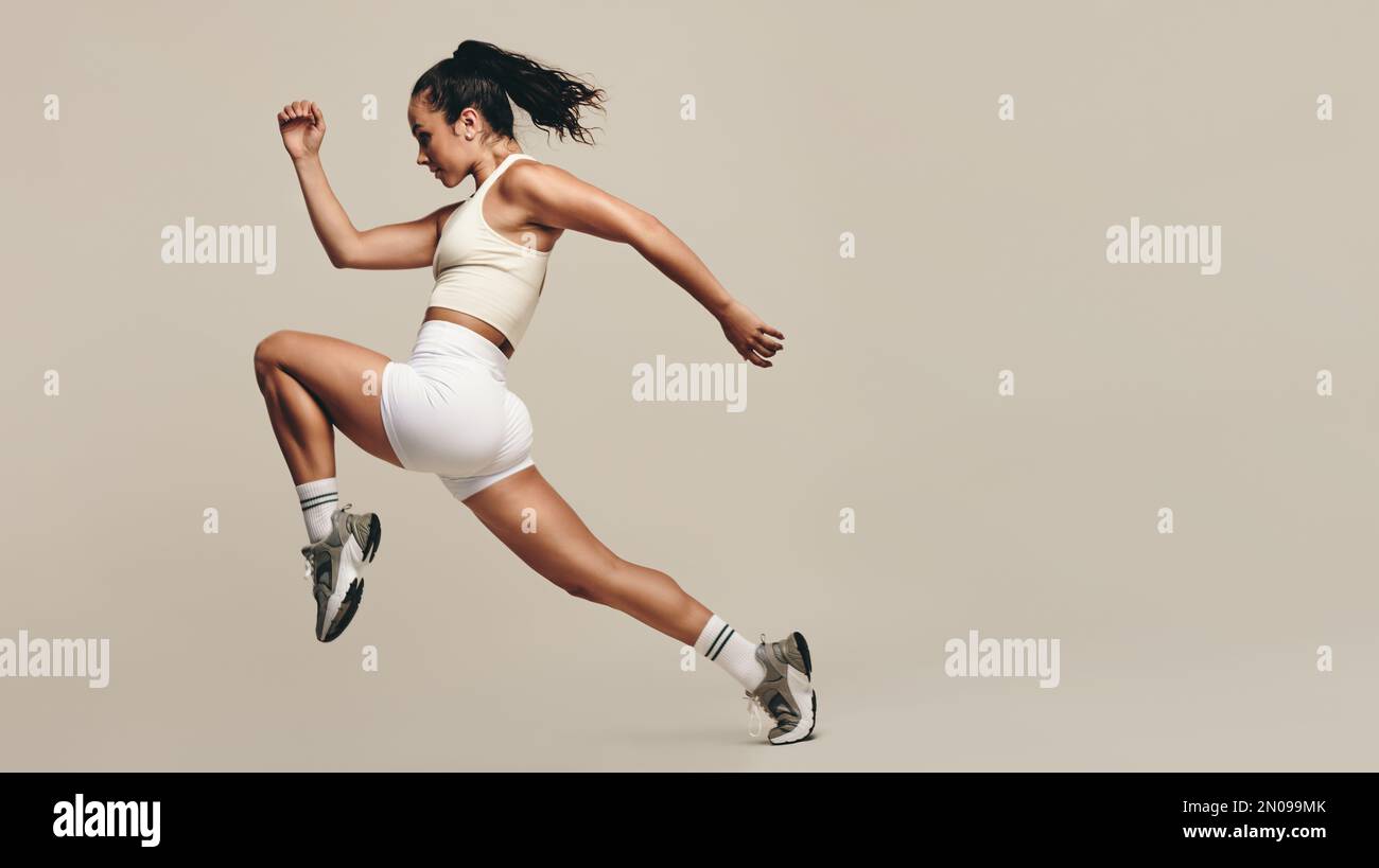 Sportswoman jumping forward, running in a vigorous studio workout. Wearing sportswear, she combines strength training with cardio exercises to improve Stock Photo