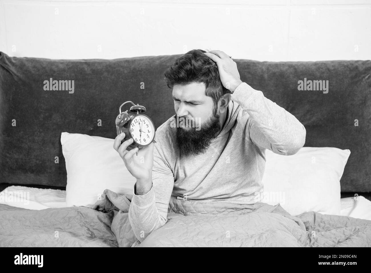Perplexed guy scratching head holding alarm clock being in bed in