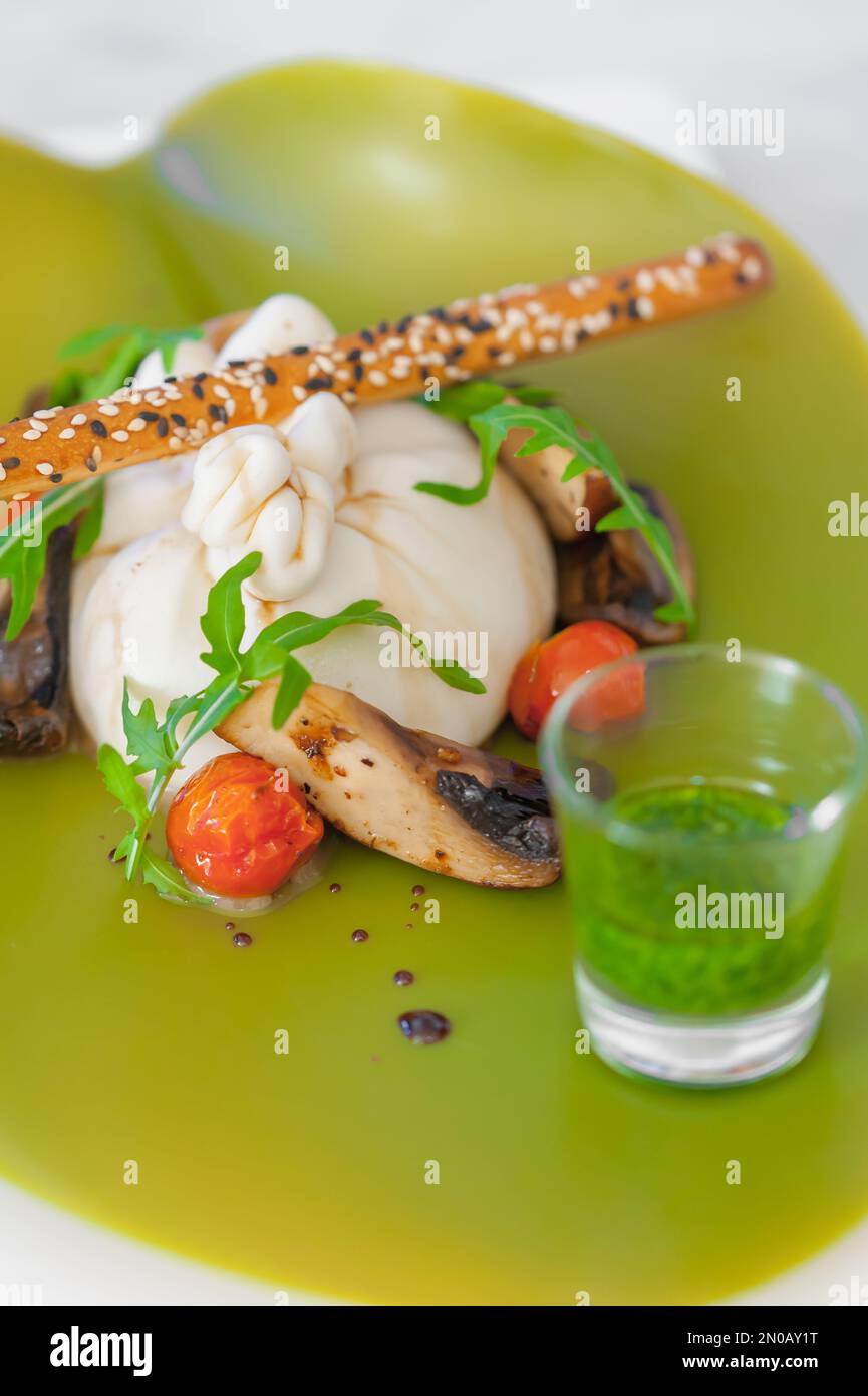 Close-up of burrata cheese salad with mushrooms, cherry tomatoes, arugula, a bread stick with sesame seeds, salsa verde. Meal on a neon green plate. Stock Photo