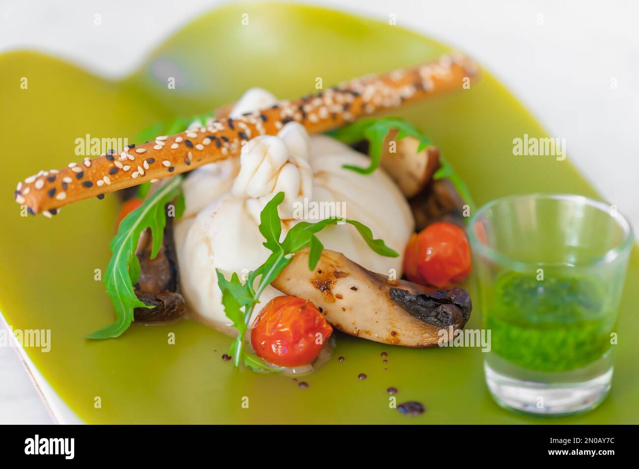 Close-up of burrata cheese salad with mushrooms, cherry tomatoes, arugula, a bread stick with sesame seeds, salsa verde. Meal on a neon green plate. Stock Photo