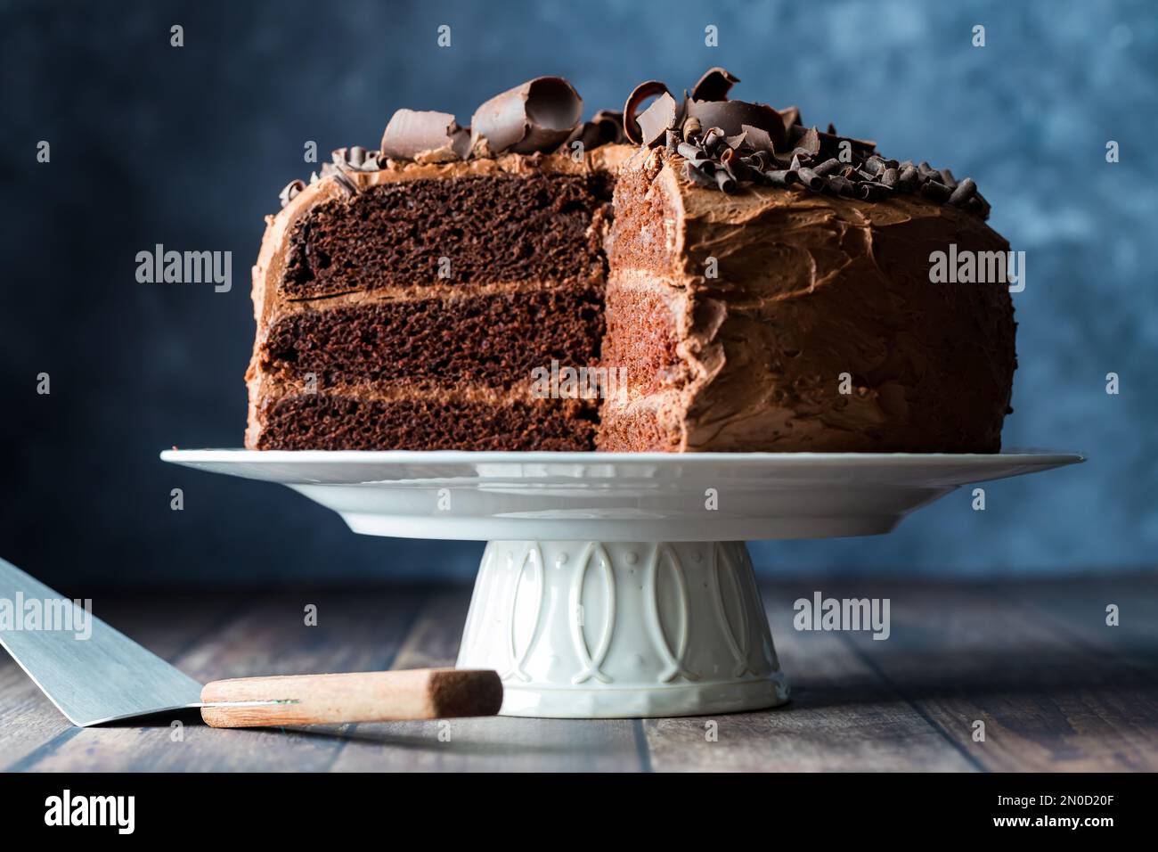 A triple layered homemade chocolate cake on a pedestal stand with slices removed Stock Photo