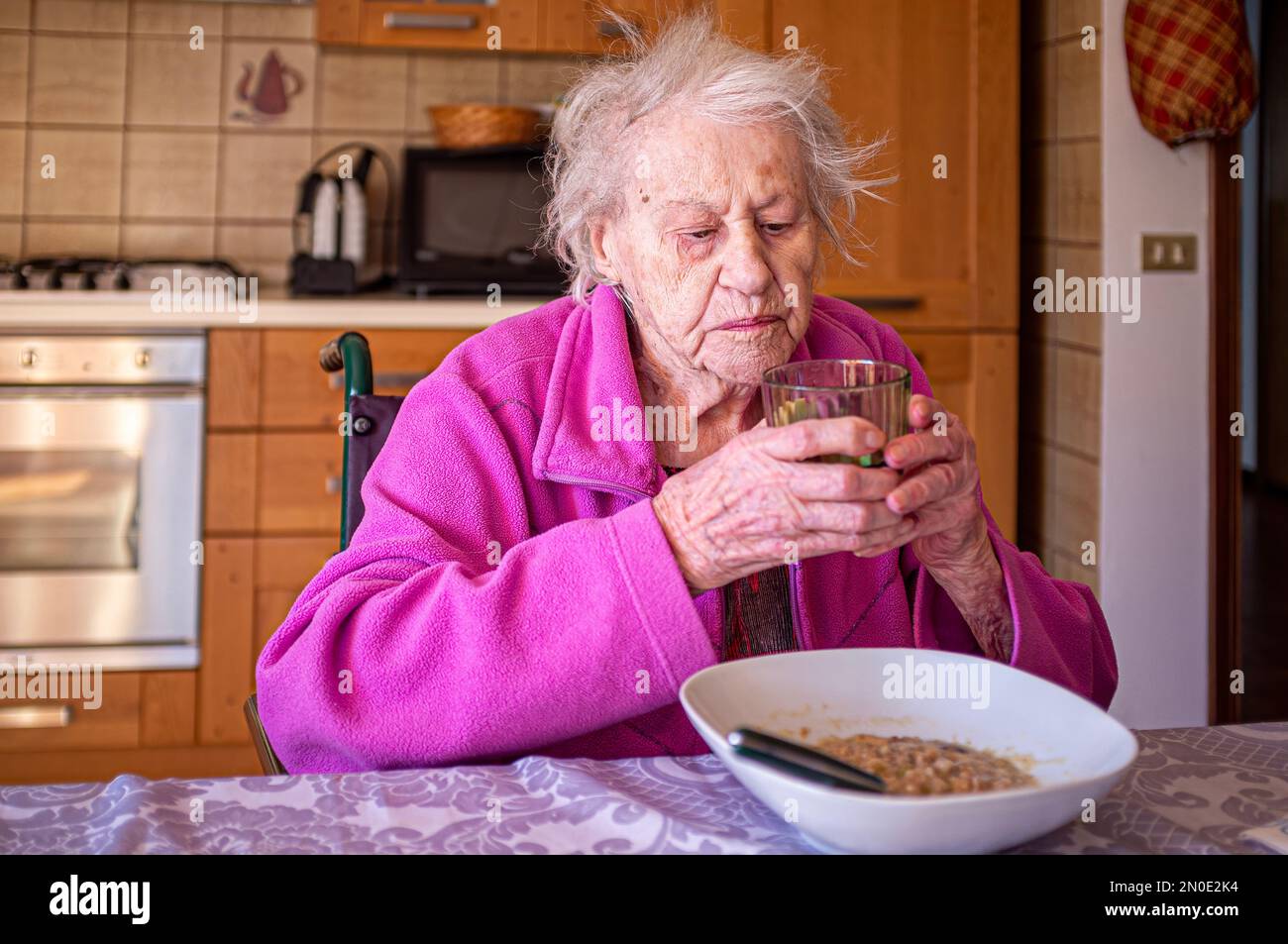 Old lady in a wheelchair with a glass of wine in hand eating alone in her kitchen Stock Photo