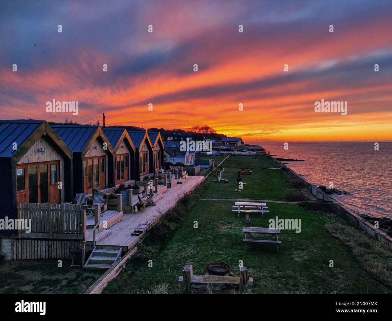 a row of same similar small tiny beachside living holiday homes on beach coast dramatic sunset reflecting building apex roof line Isle of Wight Stock Photo