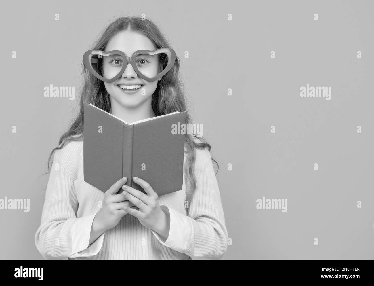 Happy kid smiling in big funny heartshaped glasses holding open school book blue background