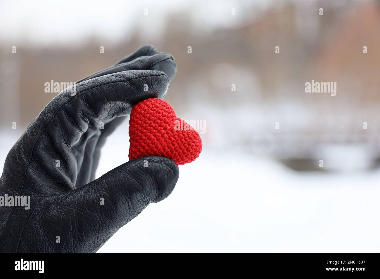 Red knitted heart in female hand in black leather glove on snow park background. Concept of romantic love, Valentine's day, winter weather Stock Photo