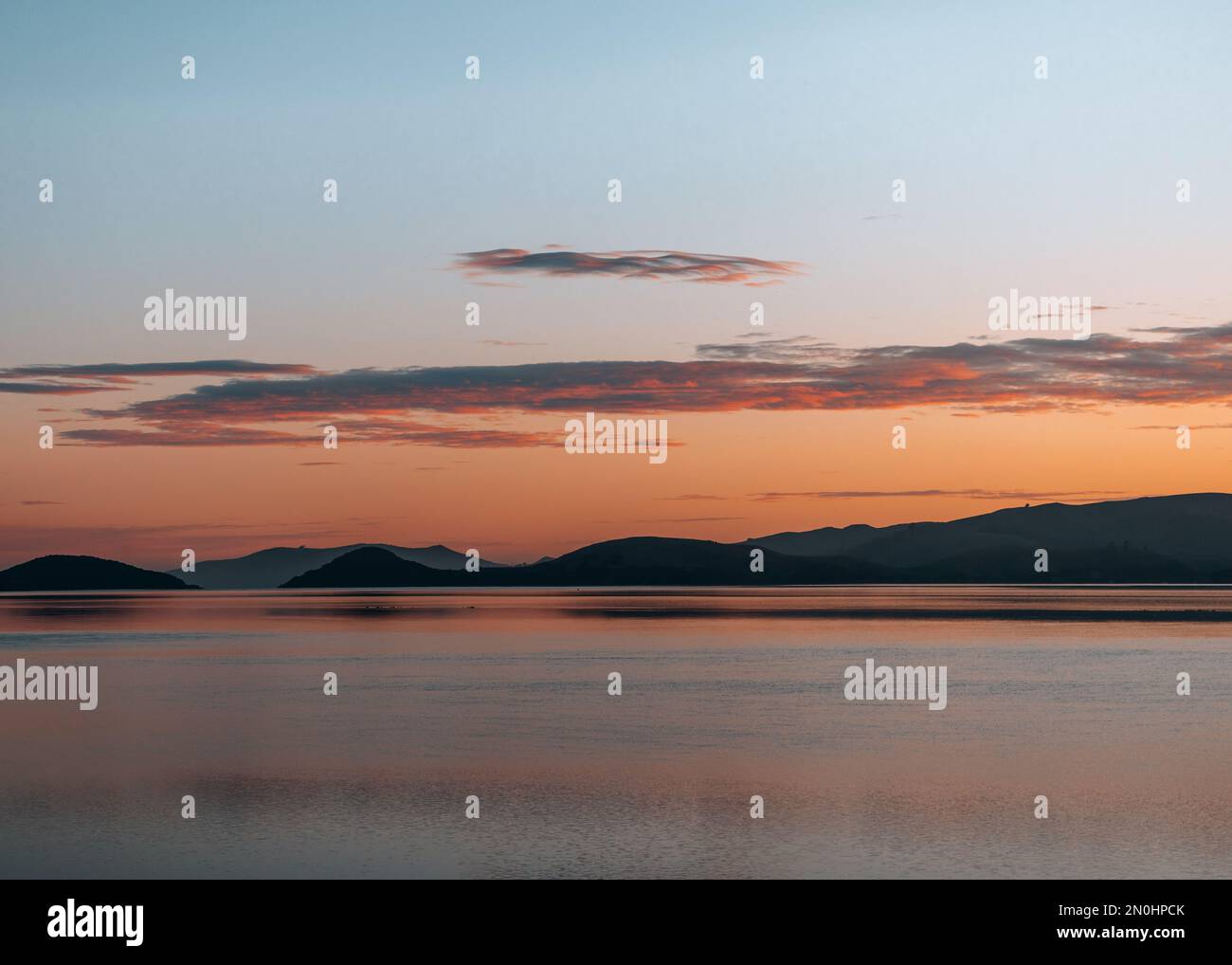 Sunrise colours and cotton candy clouds over an inlet near Otago Peninsula, New Zealand, with views of hills in the background. Stock Photo