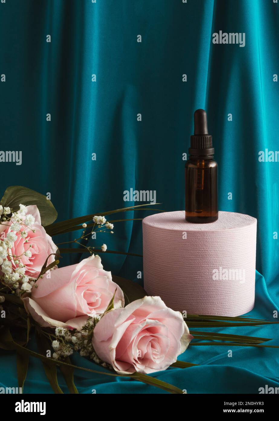 Amber glass dropper bottle on a podium with pink rose flowers. Blue silk curtain background. Skincare products, natural cosmetic. Beauty concept. Vale Stock Photo