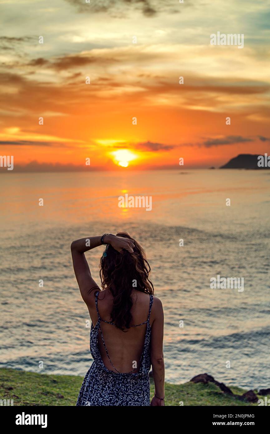 Woman with her back to the camera, watching the sun set in the sea ...