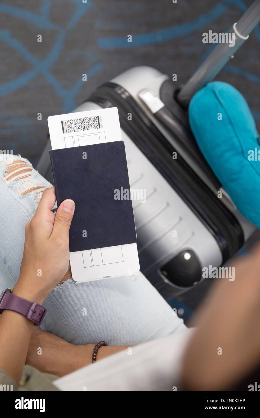 Hand holds a passport with a plane ticket inside. She is seated with a carry on next to her. Airport Stock Photo