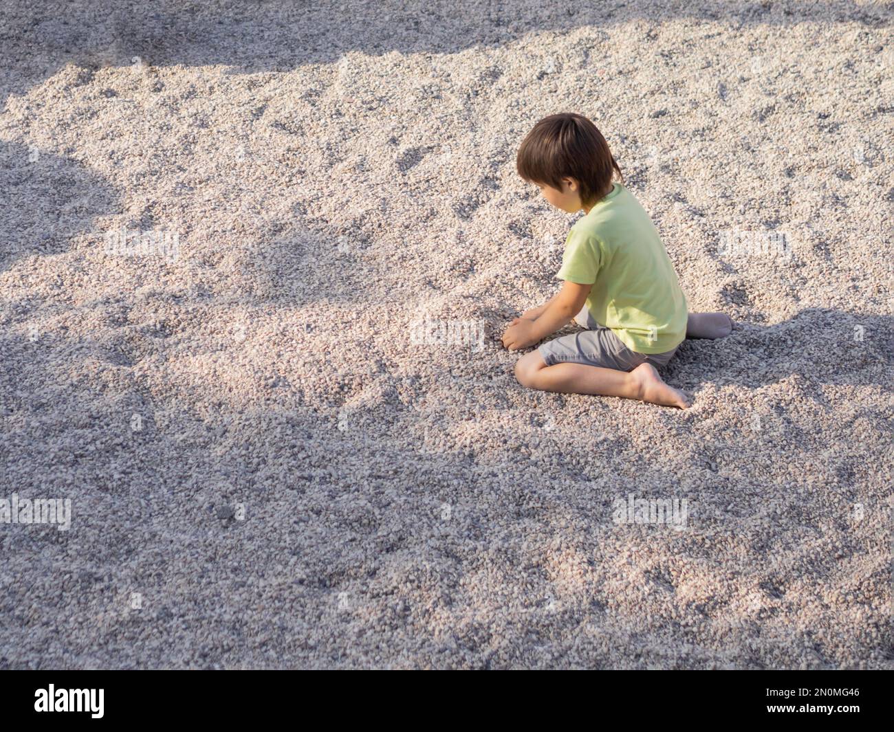 Boy is playing with gravel on modern children's playground. Developing fine motor skills and sensoric perception. Active leisure outdoors. Stock Photo