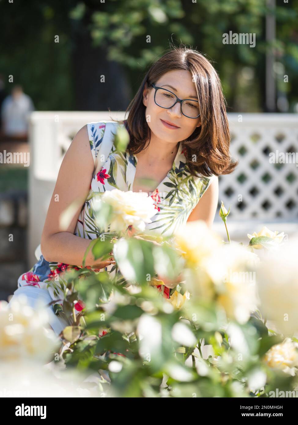 Caucasian woman is admiring of blooming roses in public park. Summer vibes. Tropical plants and flowers in bloom in garden. Stock Photo