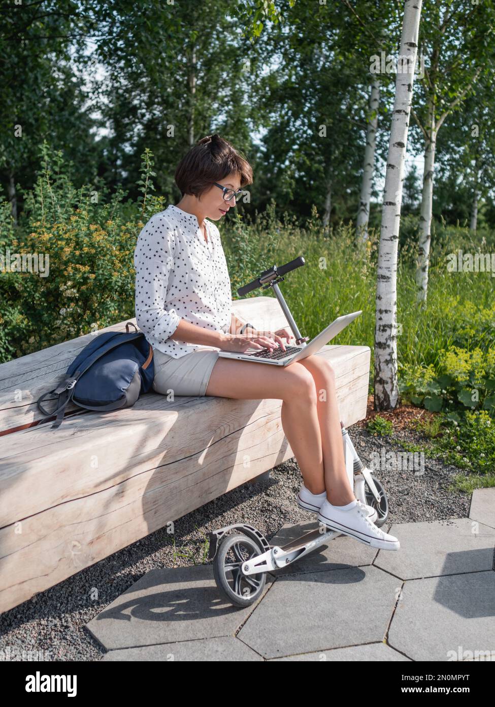 Mindful woman works with laptop on wooden bench in urban park. Summer vibes. Modern workplace for freelancers. Student educates online from outdoors. Stock Photo