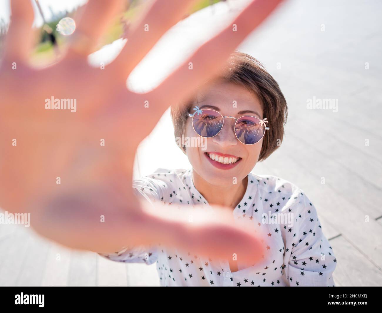 Wind ruffles short hair of freckled woman in colorful sunglasses. Smiling woman at open wooden scene of urban park. Summer vibes. Sincere emotions. Stock Photo