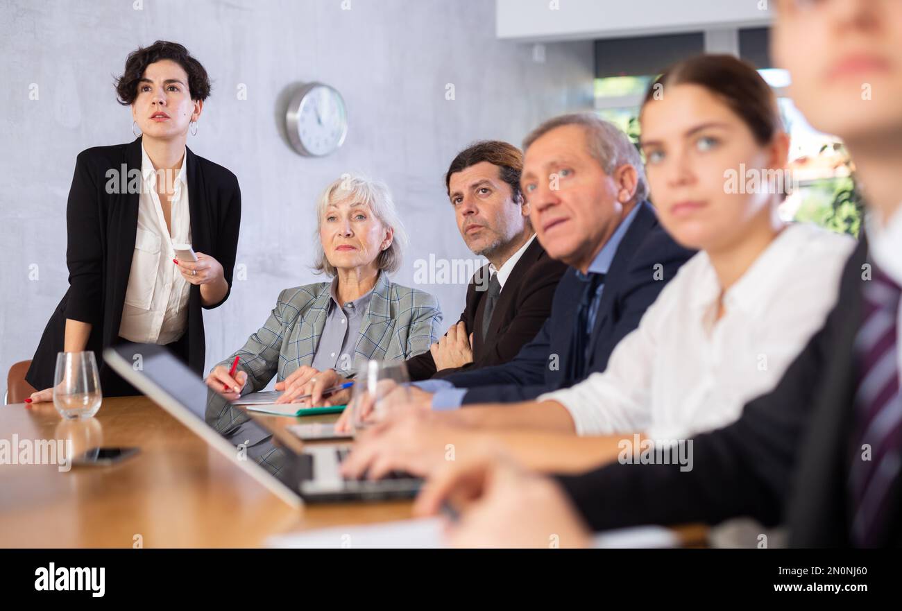 Young Latin female turning on presentation on projector while office coworkers of different ages sitting at table with laptops and notes Stock Photo