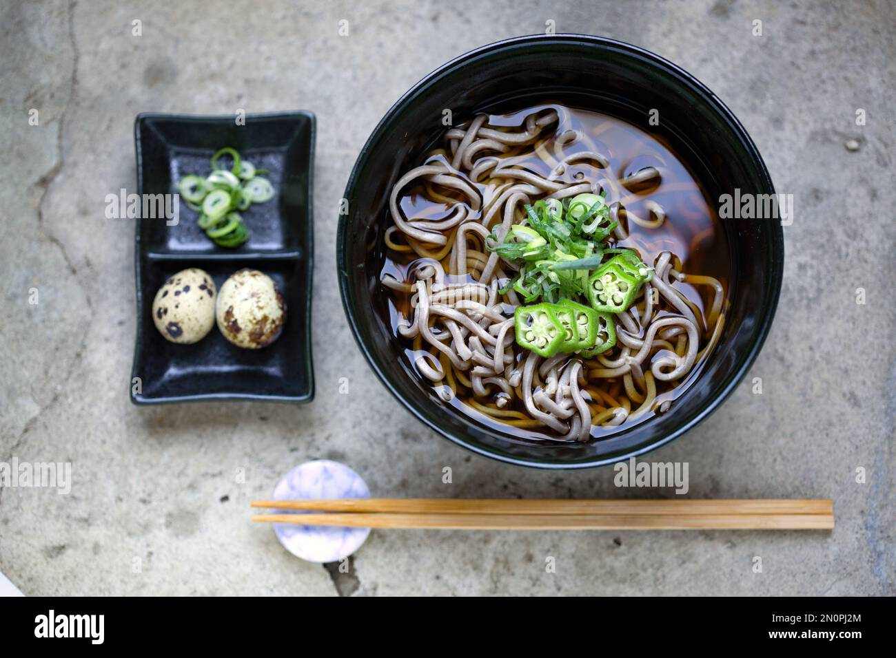A black china bowl of noodles and broth, sliced green chilli and a dish with quail's eggs and sliced vegetables. Stock Photo