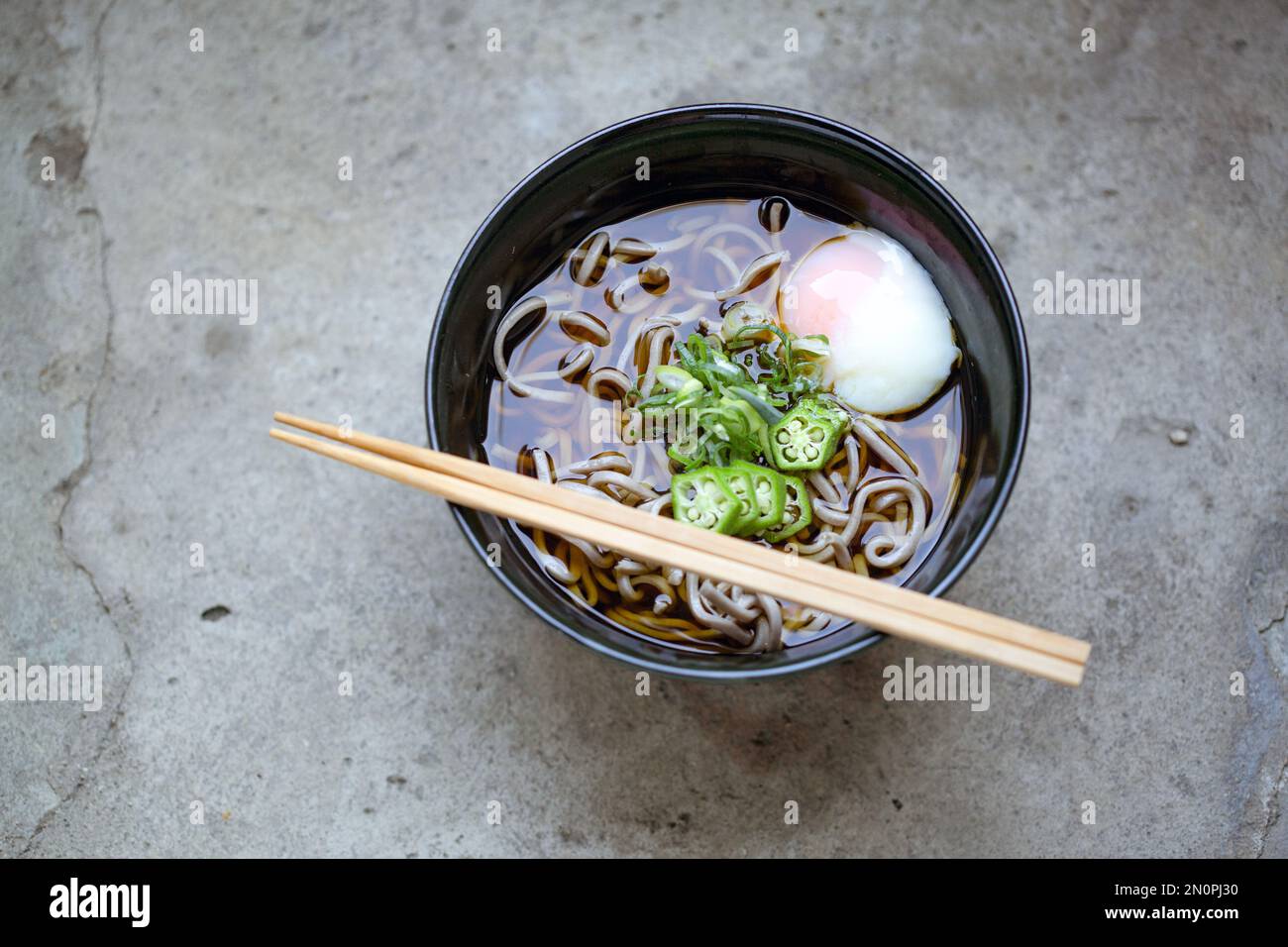 A black china bowl of noodles and broth with sliced chillis and an egg. Stock Photo